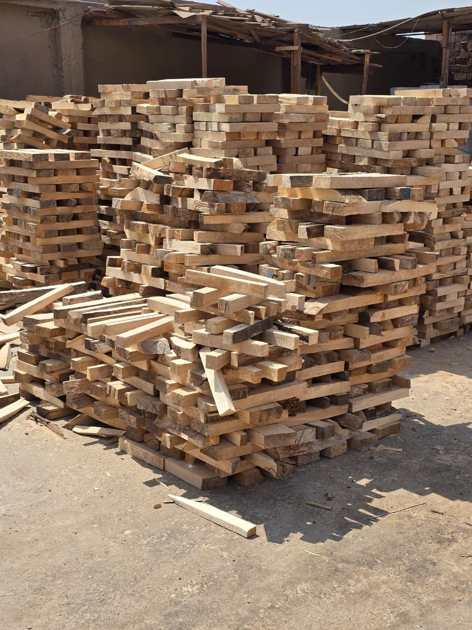 Stacks of raw lumber and wood planks piled outdoors in a warehouse or yard under a rustic roof.
